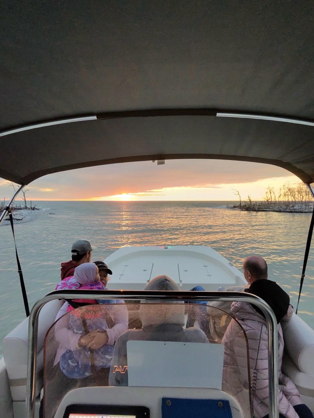 A family gathered at the bow of the pontoon under the bimini top, watching the sun set over the water